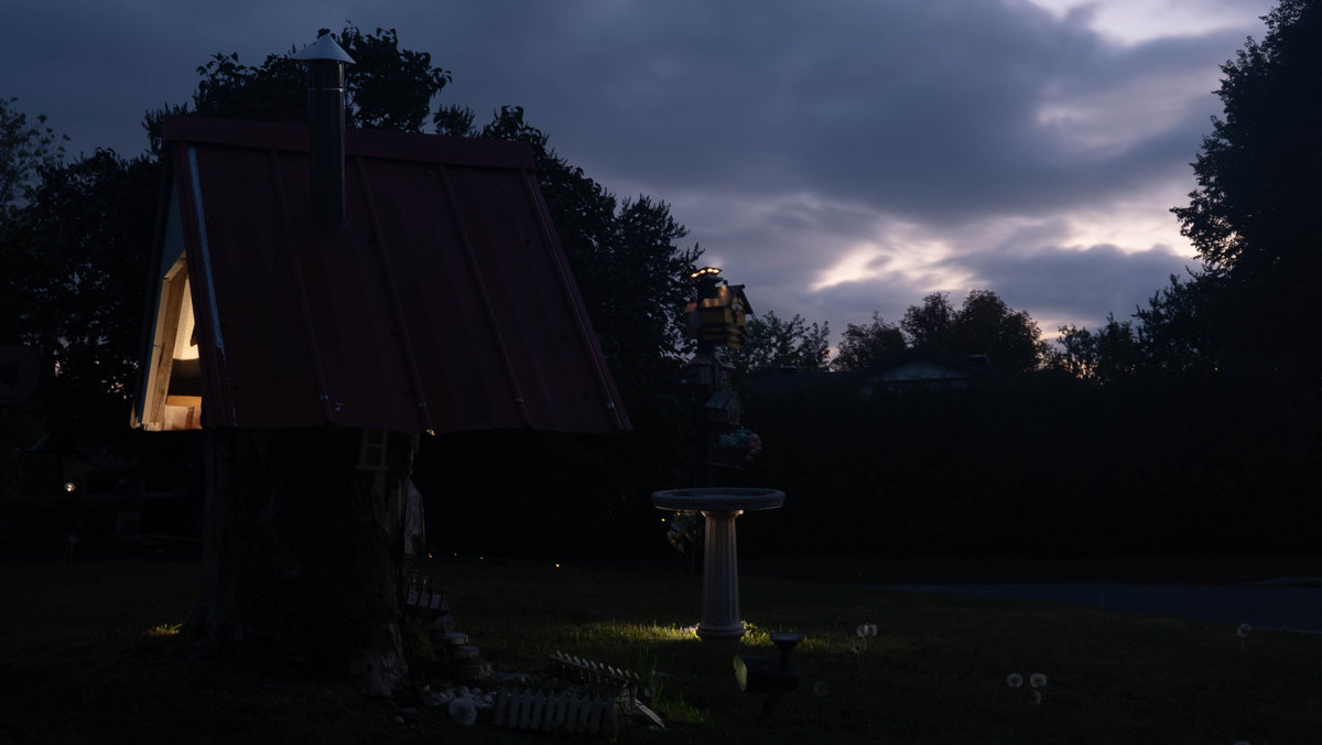 The tree house after sunset, the window and the birdbath lit by small lights under a dark blue sky