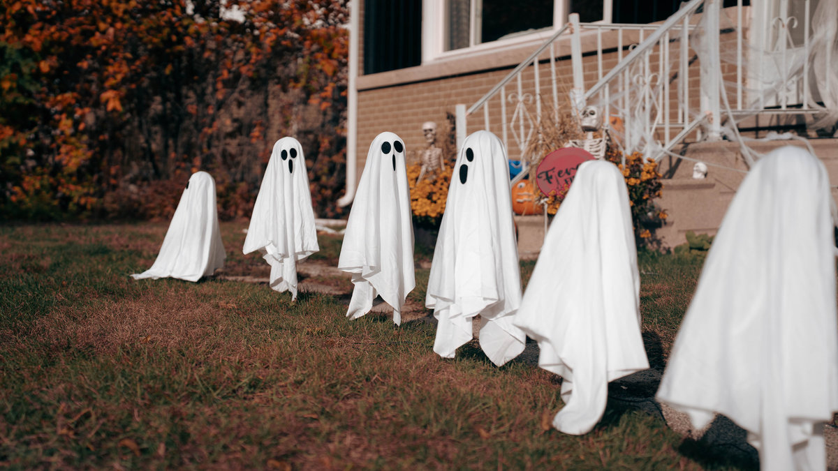 A row of white sheet ghosts on a lawn, leading toward a brick house with Halloween decorations on the steps